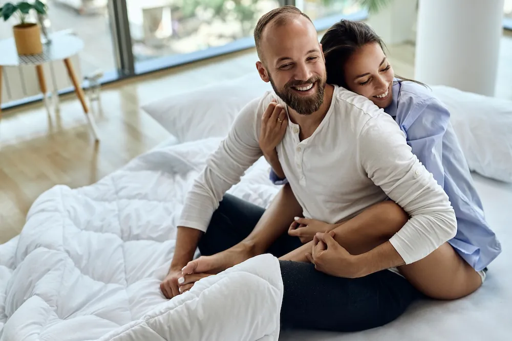 A couple happily sitting on a bed, smiling and enjoying each other's company.